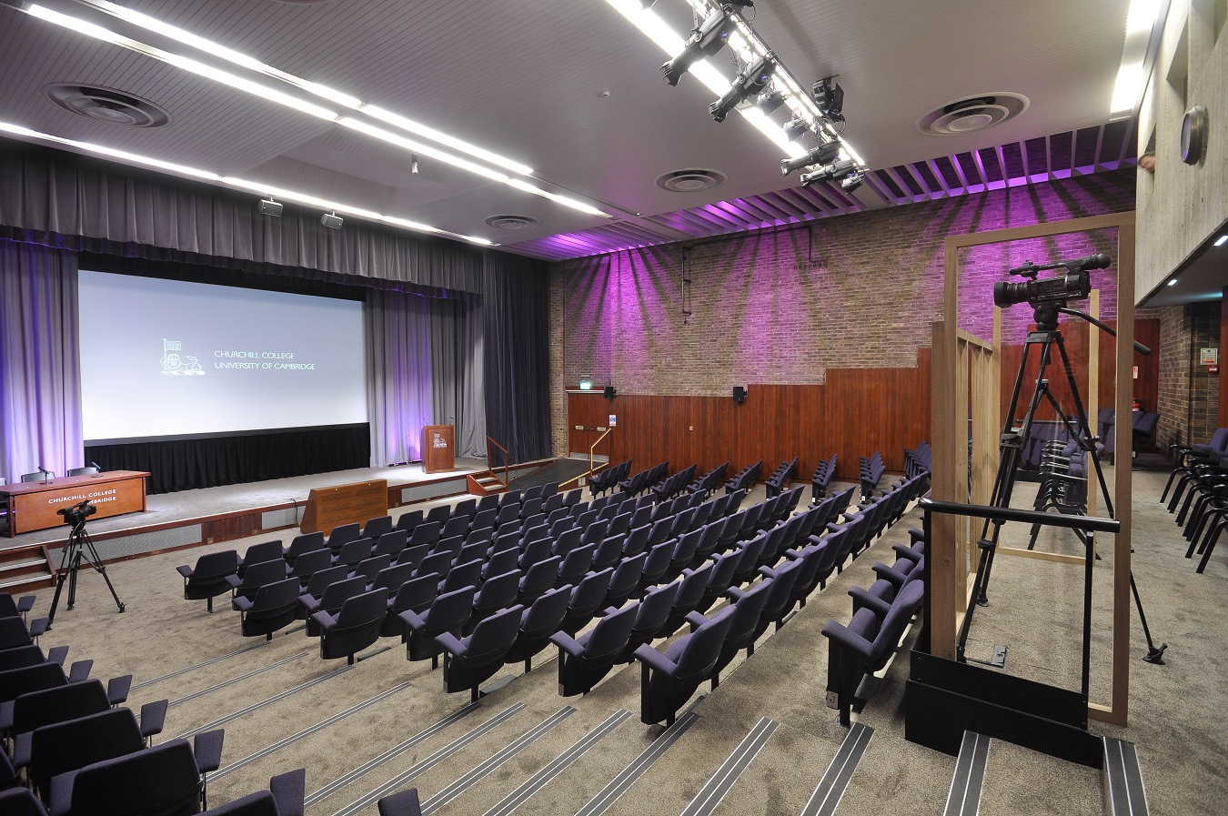 Empty auditorium with rows of navy blue seats facing a large screen. Wooden podiums are on either side of the screen, and a camera is set up on a platform to the right.