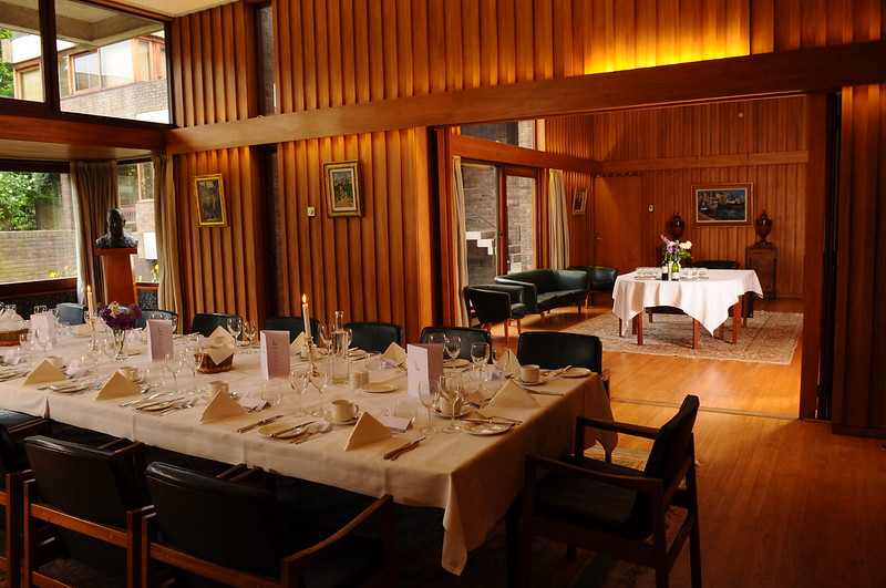 Elegant dining room with wooden paneling, featuring a long table set with white linens, glassware, and folded napkins. In the background is a cozy seating area with sofas and a coffee table on a rug. Soft lighting creates a warm ambiance.