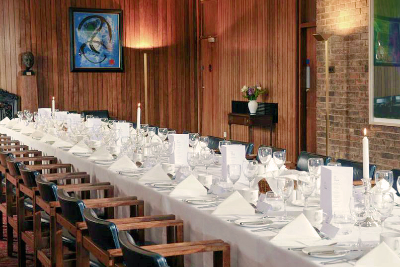 A long dining table set for a formal event. It features white tablecloths, neatly arranged glassware, silverware, plates, and folded napkins. Several chairs are lined up along each side. The room has wooden paneling and framed artwork on the walls.