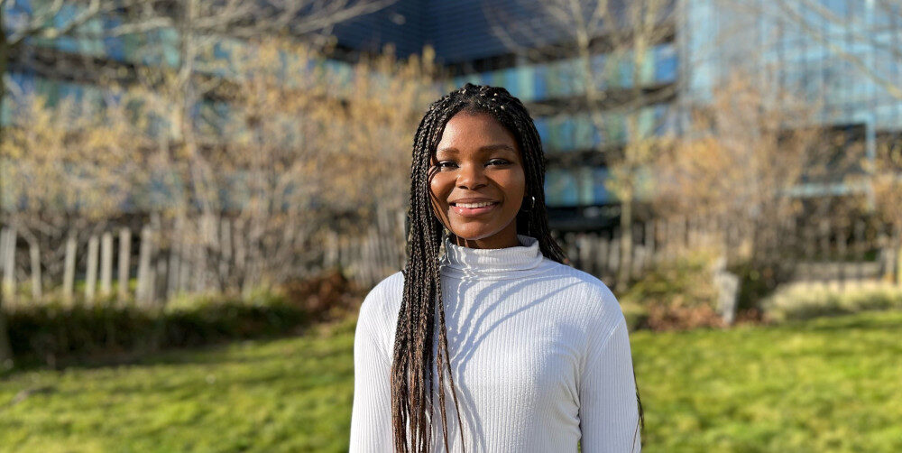 A person with long braided hair wearing a white turtleneck sweater stands smiling in a sunlit garden. The background features trees and a modern building reflecting daylight.