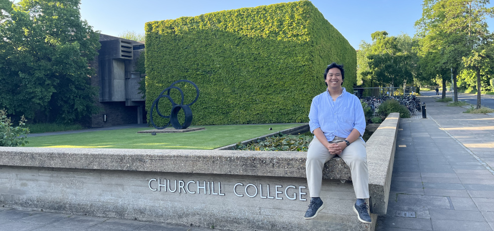 A person is sitting on a concrete wall with Churchill College written on it. Behind is a large building covered with green ivy and a circular metal sculpture on the lawn. The sky is clear and blue.