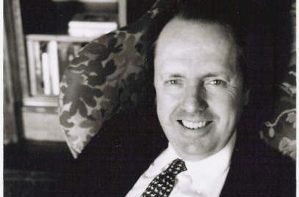 Smiling person in a suit and patterned tie, sitting in a patterned armchair. A bookshelf is visible in the background. The photo is in black and white.