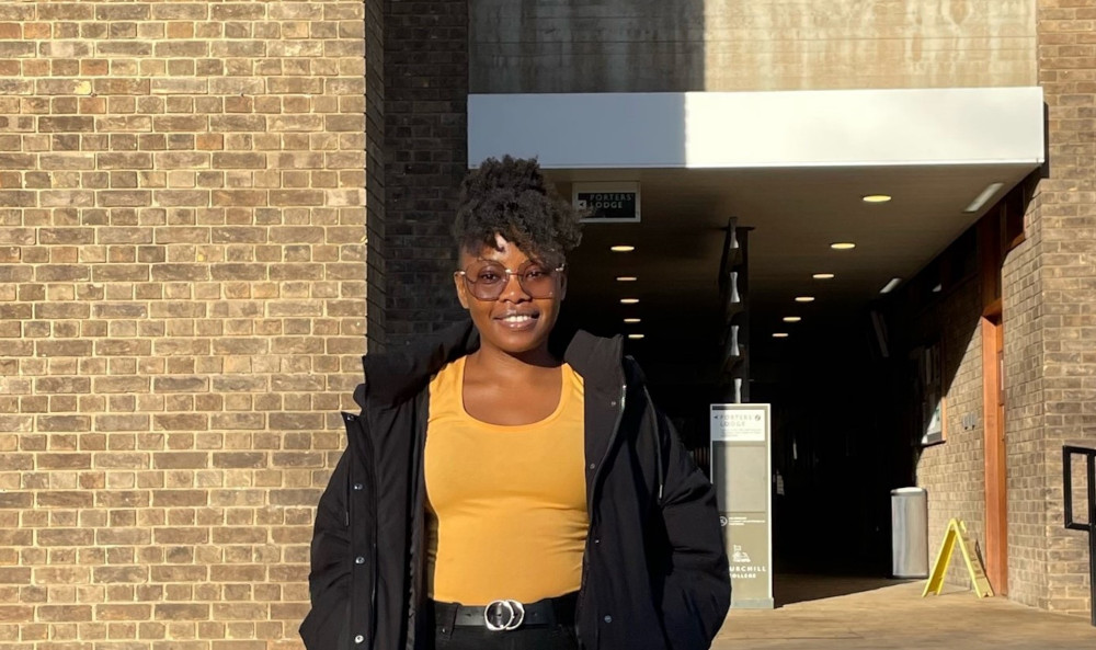 A person stands smiling outside a building with brick walls, wearing sunglasses, a yellow top, black jacket, and pants. The entrance behind them is well-lit with a sign overhead. The sun casts a shadow, highlighting their features.