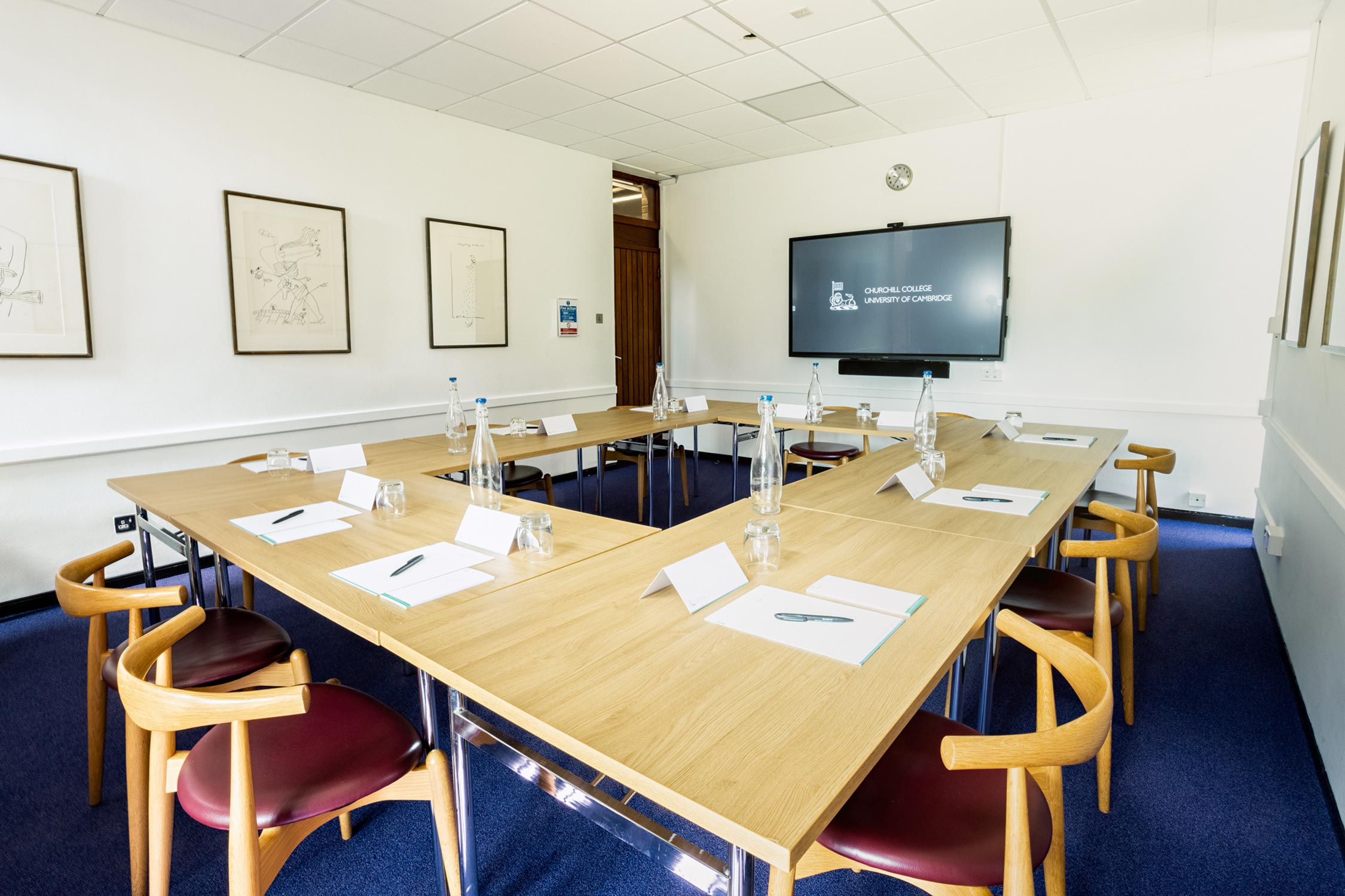 A conference room with a U-shaped setup of wooden tables. Each place has a chair, paper, pen, water bottle, and glass. Framed sketches hang on the white walls. Blue carpet covers the floor.