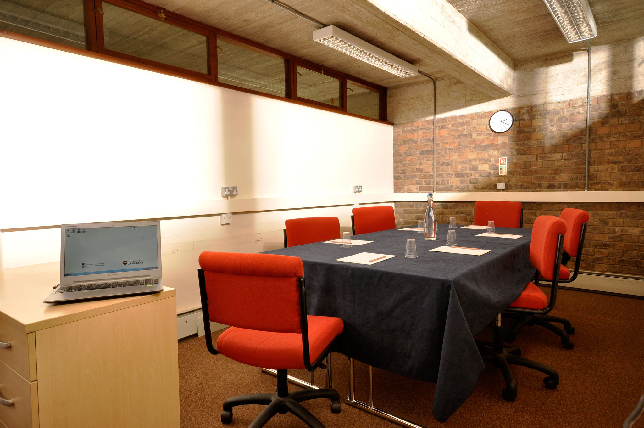 A conference room with a rectangular table covered in a black cloth. Six red chairs surround the table, which has notepads and pens. A laptop is open on a side table. The room has a brick wall, a clock, and fluorescent lighting.