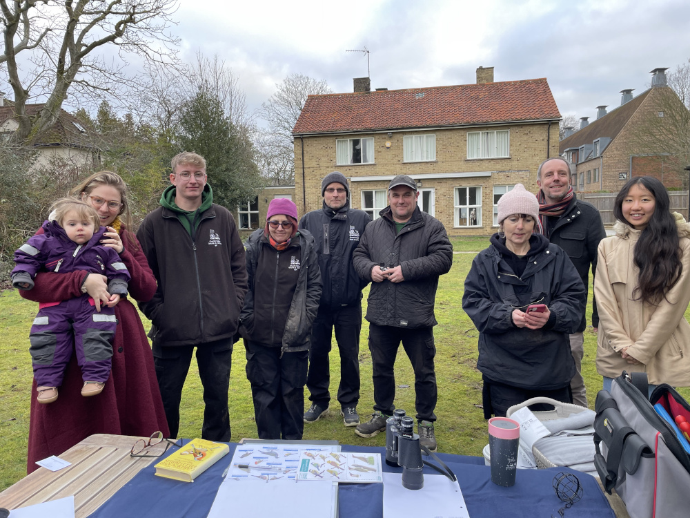 A diverse group of nine people stand outdoors on a grassy area. Some wear winter clothing. A woman holds a toddler. A table in the foreground has bird-watching equipment, maps, and books. A residential building is visible in the background.
